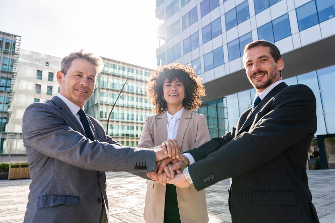 Three businesspeople uniting hands in front of building.